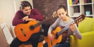 teacher guitar lessons for kids, guitar classes for kids, guitar coach for kids, The image features a girl engaged in guitar lessons with her teacher in a classroom setting in Hurstville, Sydney, Australia. The girl sits attentively, holding a guitar while her teacher stands beside her, providing guidance and instruction. The classroom is equipped with music stands, guitars, and other musical instruments, creating an engaging environment for learning. The girl's focused expression and posture demonstrate her commitment and enthusiasm for mastering the guitar. This image captures a moment of musical education and skill development as the girl embraces guitar lessons in Hurstville, Sydney, Australia.