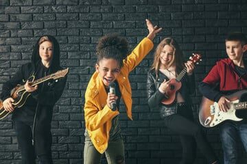 Two boys and two girls showcasing their musical talents on a stage. One girl takes center stage, confidently singing into a microphone, while the other girl and the boys accompany her on guitars, strumming in harmony. The performers exhibit a strong stage presence and skillful musicianship, reflecting the results of their dedicated music lessons. The audience's applause and the vibrant atmosphere add to the excitement and energy of the performance, as the children proudly demonstrate the culmination of their hard work and passion for music