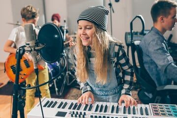 A girl, accompanied by a music teacher, showcasing her singing and keyboard skills in a vibrant classroom setting. The girl stands confidently in front of a microphone, singing passionately while simultaneously playing the keyboard. Beside her, a boy exhibits his drumming prowess, keeping the beat with enthusiasm. Another boy demonstrates his guitar skills, strumming chords with precision. The room is filled with the joyful sounds of music as the children proudly display the results of their music lessons. The teacher watches and guides them, providing support and encouragement throughout the performance.