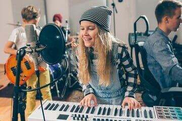 A girl, accompanied by a music teacher, showcasing her singing and keyboard skills in a vibrant classroom setting. The girl stands confidently in front of a microphone, singing passionately while simultaneously playing the keyboard. Beside her, a boy exhibits his drumming prowess, keeping the beat with enthusiasm. Another boy demonstrates his guitar skills, strumming chords with precision. The room is filled with the joyful sounds of music as the children proudly display the results of their music lessons. The teacher watches and guides them, providing support and encouragement throughout the performance.