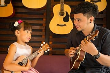 A guitar instructor in Hurstville, Sydney, guiding a young girl during a guitar lesson. The teacher is seated beside the girl, demonstrating proper finger placement on the fretboard while the girl attentively watches and learns. The room is filled with musical instruments, amplifiers, and sheet music, creating a conducive environment for learning. The girl holds a guitar, her small hands positioned on the strings, showing determination and eagerness to develop her guitar-playing skills under the guidance of her patient instructor.