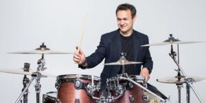 teacher singing lessons – vocal lessons, singing classes – vocal classes, singing coach – vocal coach, In a classroom in Hurstville, Sydney, Australia, a young drum teacher passionately engages in a drum lesson. The teacher is seated behind a drum set, demonstrating various drumming techniques with precision. The focus is solely on the teacher, who is dedicated to imparting knowledge and skills to the students (not visible in the image). The classroom is filled with drum sets, cymbals, and other percussion instruments, creating an immersive and energetic learning environment. This image captures the teacher's expertise and enthusiasm in providing drum lessons to aspiring musicians